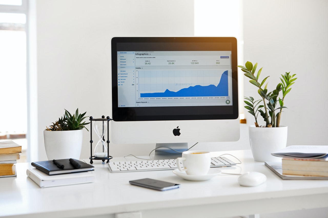 Desk with iMac displaying graph, flanked by plants, books, notebook, coffee cup, phone, hourglass, keyboard, and mouse.